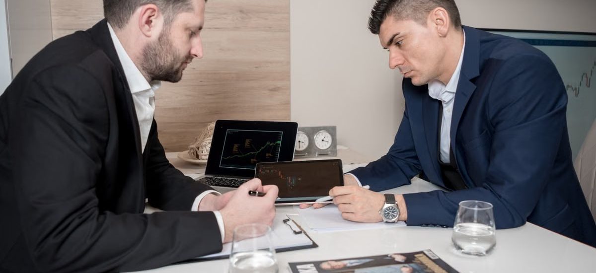 Two businessmen in suits discussing cryptocurrency trends using digital devices in an office setting.