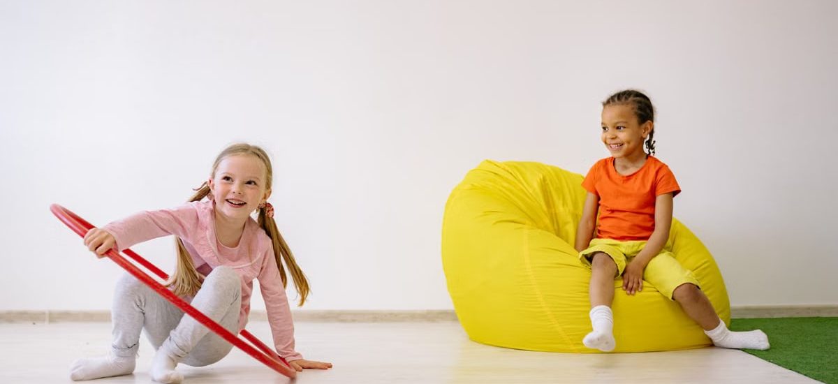 Two children enjoying playtime indoors with a hula hoop and bean bag, full of joy and fun.